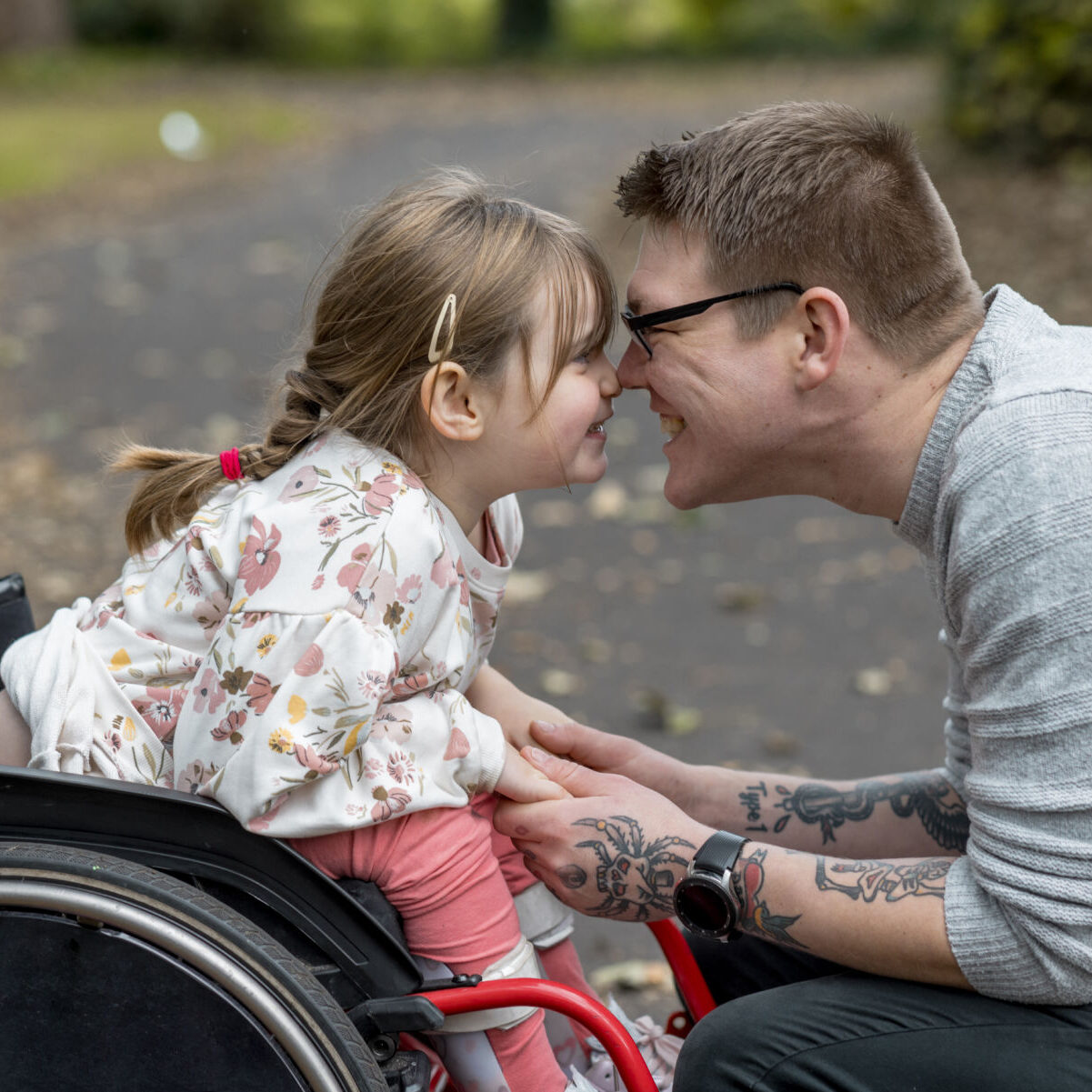 A close up side view of a father and his young daughter who is a wheelchair user having a cute affectionate moment with each other whilst on a family day out in a beautiful public park in Newcastle upon Tyne in the North East of England.